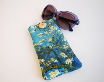 May include: A blue and green floral patterned glasses case with a wooden button closure. The case is lying on a white surface next to a pair of purple sunglasses.