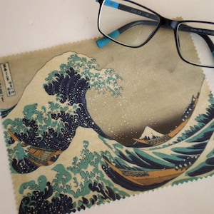 Può includere: Un panno per la pulizia in microfibra con un'immagine stampata di "La Grande Onda di Kanagawa" di Katsushika Hokusai. Il panno è beige con un design a onde blu, bianco e nero. Il testo giapponese "神奈川沖浪裏" è stampato nell'angolo in alto a sinistra.