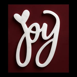 May include: A white, cursive "joy" sign with a heart-shaped dot above the "j" against a deep red background. The word is stylized and appears to be made of a glossy material. The background is a solid, matte color.