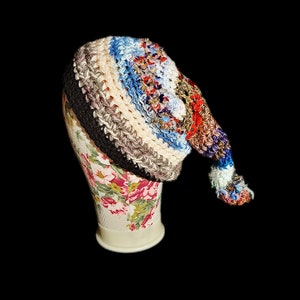 Puede incluir: Un gorro de crochet colorido con una cola larga. El gorro está hecho de hilos de diferentes colores, incluyendo negro, blanco, azul, rojo y marrón. El gorro está sentado en una cabeza de maniquí con estampado floral.