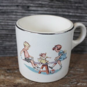 May include: White ceramic mug with a silver rim. The mug features a colorful illustration of three children playing on a seesaw. The seesaw is supported by a wooden A-frame.