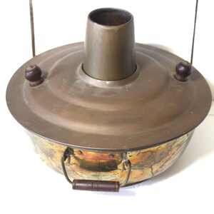 May include: Vintage copper and brass cooking pot with a central chimney and wooden handles. The pot has a round base and a lid with two wooden knobs. The pot is designed for cooking and has a rustic appearance.