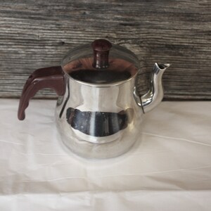May include: A silver teapot with a brown plastic handle and lid. The teapot is sitting on a white surface.