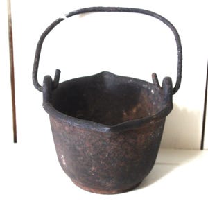 May include: A small, antique cast iron cauldron with a curved handle. The dark brown pot has a rounded body and a slightly flared rim. The handle is attached with two small loops on either side of the rim.