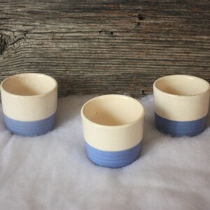 May include: Three white ceramic cups with a blue band around the bottom. The cups are sitting on a white surface.