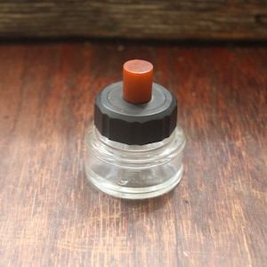 May include: A clear glass bottle with a black lid and a red top. The bottle is empty and has a textured surface.