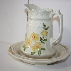 May include: A white ceramic pitcher with a floral design featuring yellow and green flowers. The pitcher is sitting on a matching white ceramic saucer.