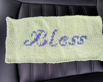 May include: A light green knitted rectangle with the word "Bless" in blue. The knitted item is resting on a black leather surface. The word "Bless" is in a cursive font.