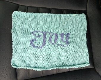 May include: A knitted square in a light teal color with the word "JOY" in a darker purple hue. The word is centrally placed and knitted into the fabric. The item is likely handmade.