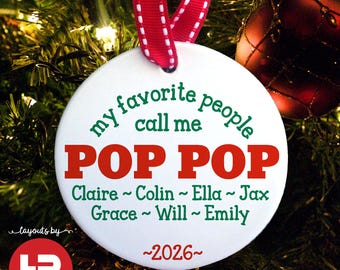 May include: A round white ceramic Christmas ornament featuring the green text my favorite people call me, the red text POP POP, and the green names Claire, Colin, Ella, Jax, Grace, Will, and Emily, with the red year 2026 and a red ribbon.