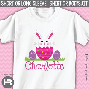 May include: White t-shirt with a pink Easter bunny peeking out of an egg with purple and pink polka dots. The bunny has a pink nose and pink ears. The text "Charlotte" is written in pink below the bunny.