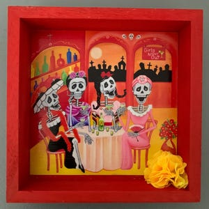 May include: A framed artwork featuring a vibrant scene of four skeleton figures at a bar, with the text "Girls Night Out." The artwork is set in a red frame and includes a yellow flower.