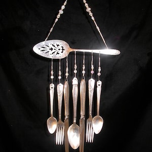 May include: A handcrafted wind chime made from repurposed silverware. A large, ornate serving spoon is suspended from a beaded strand, with smaller spoons, forks, and knives hanging below. The silverware is silver-toned, creating a unique and decorative piece.