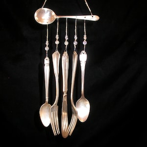 Può includere: Una carillon fatta a mano con posate riciclate. Un grande cucchiaio sospende cucchiaini, forchette e un coltello più piccoli, ciascuno collegato da fili di perline. Le posate d'argento sono appese su uno sfondo nero, creando un pezzo decorativo unico.