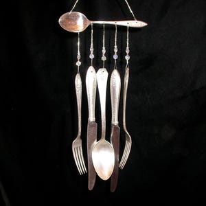 May include: A unique wind chime crafted from recycled cutlery. A large spoon is at the top, with forks, knives, and spoons hanging below. Clear and blue beads provide decoration. The background is a plain black.