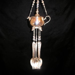 May include: A silver-coloured wind chime featuring a small teapot suspended from beaded strands. Below the teapot hang spoons and forks. The wind chime is designed with intricate details and hangs against a black background.