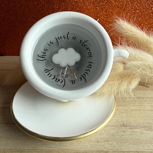 Könnte beinhalten: Weißes Tee-Set mit Untertasse und Tasse, goldfarbener Rand. Die Tasse zeigt ein Wolken- und Bergdesign mit dem Text "This is just a storm inside a teacup..." in schwarzer Schrift. Der Hintergrund ist ein glitzerndes Orange.