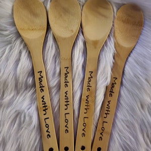 May include: Four bamboo cooking spoons with the words "Made with Love" written in black ink on the handles. The spoons are light brown and rest on a fluffy, white surface.