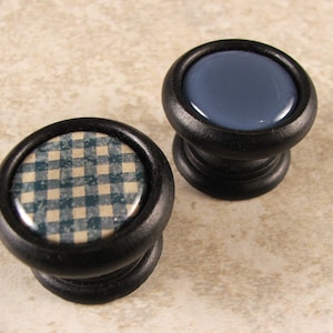 May include: Two black wooden knobs with different patterned tops. One knob has a blue and white gingham pattern, and the other has a solid blue top.