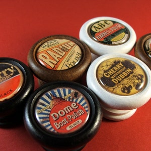 May include: Six vintage shoe polish tins with various labels. The tins are round and have a wooden base. The labels feature different colors and designs, including a cherry blossom, a dome, a radiating sun, a horse and carriage, and a dubbin.