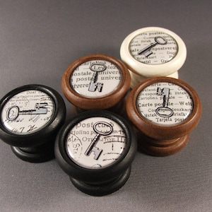 May include: Four wooden knobs with different colored bases and vintage key designs on top. The designs feature a key on a background of old postage stamps and text. The knobs are in black, brown, white, and brown.