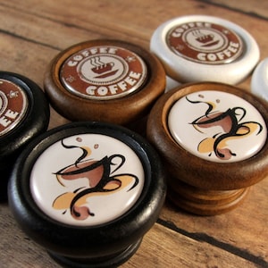 May include: Five wooden knobs with a coffee cup design. The knobs are in black, white, and brown wood. The coffee cup designs are in brown, white, and black.