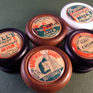 May include: Six vintage milk bottle lid knobs with wooden bases. The lids feature colourful labels with text such as "Gill's Fresh Pasteurised Milk", "Fawndale Milk", "Premier Pasteurised Milk", "Hall's Dairy Pasteurised Table Cream", and "Hartwig Dairy Buttermilk".