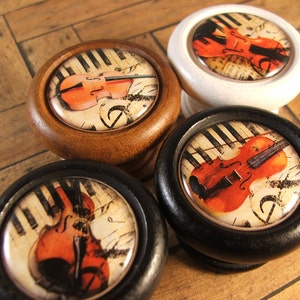 May include: Four wooden knobs featuring a violin and musical notes design. The knobs are in different colours: brown, white, black, and black.
