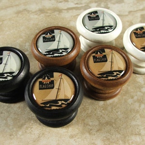May include: Six wooden cabinet knobs with a nautical theme. Each knob features a white sailboat with a red flag that says "Flagship" on a brown, black, or white background.