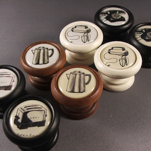 May include: Set of eight kitchen-themed cabinet knobs. The knobs are made of wood and have a variety of designs, including a toaster, a coffee pot, a mixer, and a telephone. The knobs are in black, white, and brown.