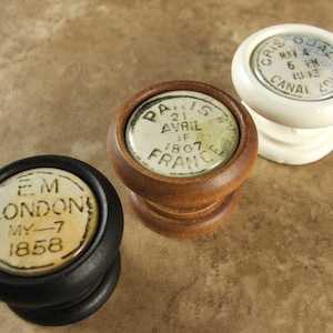 May include: Three vintage wooden knob pulls with ceramic tops. The top of the left knob is black with white text that reads "EM LONDON MY-7 1858". The middle knob is brown with white text that reads "P 21 AVRIL F 1807 FRANCE". The right knob is white with black text that reads "CRIS OSAL MAY 4 6 KM 1913 CANAL ZONE".