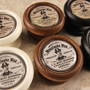 May include: Five wooden knobs with silver labels that read "Moustache Wax" in a vintage font. The labels feature a man with a mustache wearing a top hat. The knobs are in various colors, including white, black, and brown.