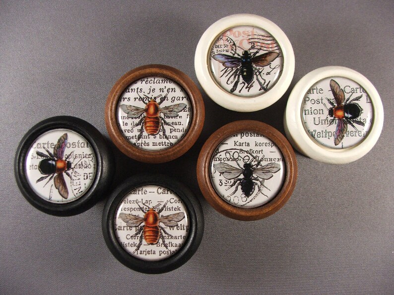Op de afbeelding: Zes ronde houten en witte knoppen met zwarte en bruine bijenillustraties en vintage tekst. De tekst bevat woorden als "Carte Postale", "Karta koresp", "Post", "Union", "Weltpost" en "r&eacute;clames".