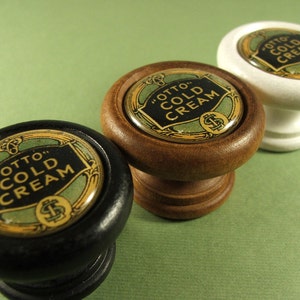 May include: Three vintage cabinet knobs with a wood, white, and black finish. Each knob features a round, green and gold label with the text "Otto's Cold Cream".