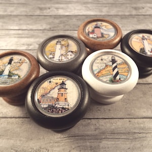 May include: Six different wooden knobs with images of lighthouses on them. The knobs are painted in various colors, including black, white, brown, and gray. Each knob features a different lighthouse design.