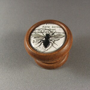 Op de afbeelding: Een houten knop met een ronde glazen bovenkant met een zwart-witte afbeelding van een bij met tekst in een vreemde taal.