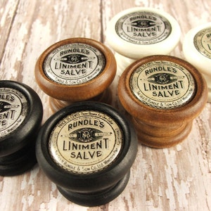 May include: Five wooden containers with silver labels that read "RUNDLE'S LINIMENT SALVE" in a vintage style font. The containers are in various colors, including black, brown, and white.
