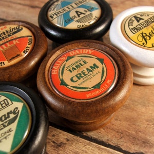 May include: A collection of vintage wooden knobs with colorful, aged labels. The labels feature text for various dairies, including "Cliff Herring Dairy," "Pruett Bros," and "Beck's Dairy." The knobs are in various colors, including black, white, and natural wood.