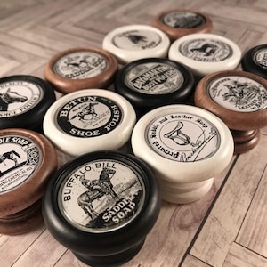 May include: A collection of vintage-style wooden knobs with black and white labels featuring different shoe and saddle polishes. The labels include text such as "Fiebing's Saddle Soap", "Betun Shoe Polish", "Buffalo Bill Saddle Soap", and "Day Martin Boot Polish".