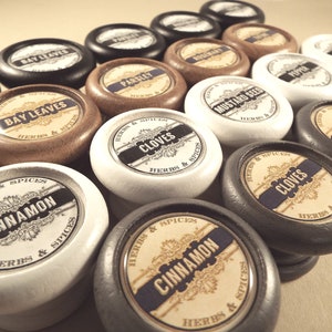 Peut inclure: Une collection de pots à épices ronds en bois avec des couvercles noirs, blancs et bruns. Chaque couvercle porte une étiquette avec le nom d'une épice différente, comme "Cannelle", "Clous de girofle" et "Graines de moutarde".