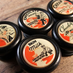 Peut inclure: Six boutons de cabinet noirs et blancs avec des publicités vintage pour le lait. Les boutons présentent des illustrations de laitiers livrant du lait, avec du texte qui lit "Homogénéisé Vitamine D Lait Sealon Hood Protégé", "Boisson au chocolat Sealon Hood Protection", "Chocolat au lait Sealon Hood Protection", "Lait écrémé Sealon Hood Protégé", et "Crème Sealon Hood Protégé".