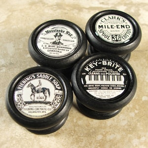 May include: Four black wooden knobs with vintage advertising labels. The labels feature products like saddle soap, moustache wax, spool cotton, and piano key cleaner.