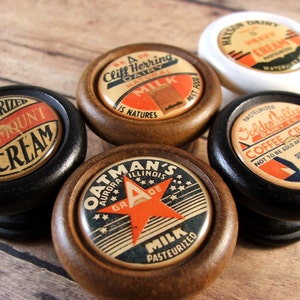 May include: Five vintage wooden knobs with different labels. The labels are for dairy products, including cream, milk, and coffee cream. The labels are in various colors, including red, white, blue, and black.