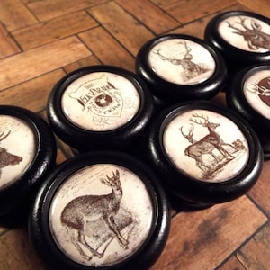 May include: Set of seven black and white deer themed cabinet knobs. Each knob features a different deer design, including a stag, a doe, and a fawn. The knobs are made of wood and have a black finish.