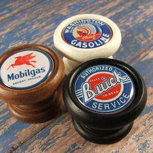 May include: Three vintage-style cabinet knobs with different designs. The first knob is brown wood with a white and red Mobigas logo. The second knob is white with a red, white, and blue Washington Chief Gasoline logo. The third knob is black with a blue and red Buick Authorized Service logo.