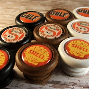 May include: Eight vintage-style wooden knobs featuring different gas and oil company logos. The logos include Gulf, Shell, and Standard Oil. The knobs are in various colours, including black, white, and brown.