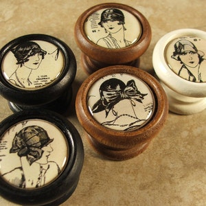 May include: Five vintage-style cabinet knobs with wooden bases and round, white ceramic tops featuring black and white illustrations of women wearing hats. The illustrations are from a vintage fashion magazine.