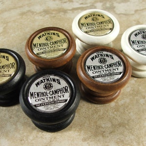 May include: Five round wooden knobs with a gold and black label that reads "FREE WATKINS MENTHOL-CAMPHOR OINTMENT NET WT. 1/8 OZ THE J.R. WATKINS COMPANY". The knobs are in various colors, including black, brown, and white.