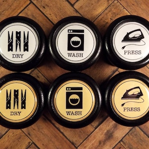 May include: Six black knobs with white and gold labels. The labels have laundry symbols and text. The symbols are for washing, drying, and pressing clothes. The text on the labels reads "WASH", "DRY", and "PRESS".