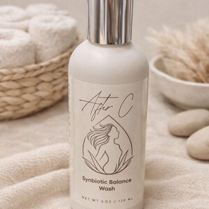 May include: A white bottle with a silver cap, labeled "After C" with a woman's silhouette. The bottle is labeled "Synbiotic Balance Wash" and contains 4 oz / 120 mL. The background includes a woven basket with rolled towels and decorative elements.